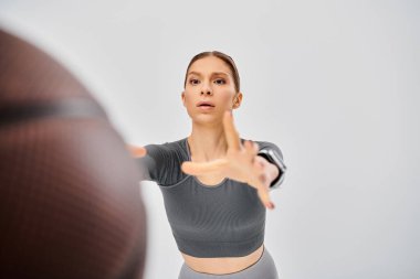 A sporty young woman in gray top effortlessly throws a basketball on a neutral background. clipart