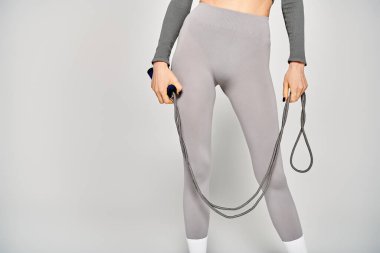 Gri pantolonlu sportif bir genç kadın, gri bir arka plana karşı atlama halatı tutuyor..