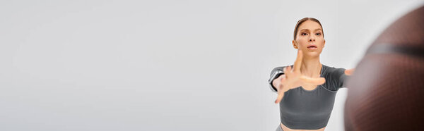 A sporty young woman in gray top effortlessly throws a basketball on a neutral background.