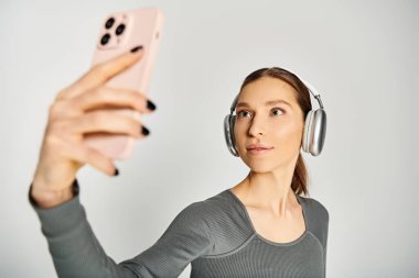 Aktif durumda olan genç bir kadın, gri bir arka planda elinde cep telefonuyla kulaklıkla müzik dinliyor..
