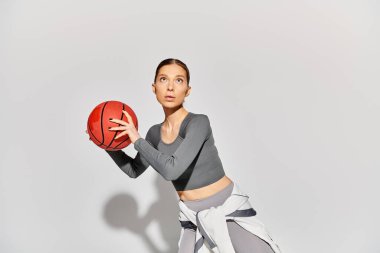 Aktif giyinmiş genç bir kadın gri bir arka plana karşı sağ elinde basketbol topu tutuyor..