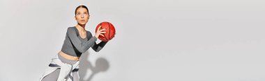 Aktif giyinmiş genç bir kadın gri arka planda kırmızı bir basketbol topu tutuyor..