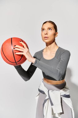Aktif giyinmiş genç bir kadın gri arka planda basketbol topunu nazikçe tutuyor..