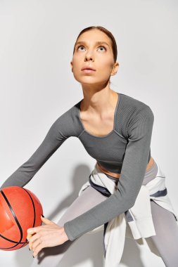 Aktif giyinmiş genç bir kadın gri bir arka plana karşı sağ elinde basketbol topu tutuyor..