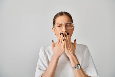 A young woman in a white t-shirt and glasses covering her mouth with her hands on a grey background. clipart