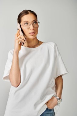 Gözlüklü şık bir genç kadın cep telefonuyla gri bir arkaplanda sohbet ediyor, nişanlı ve modern görünüyor..