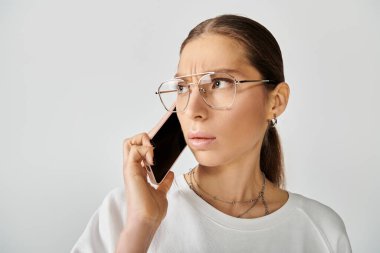 Beyaz tişörtlü ve gözlüklü genç bir kadın gri bir arka planda cep telefonuyla konuşuyor..