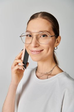 Beyaz tişörtlü ve gözlüklü genç bir kadın gri bir arka planda cep telefonuyla sohbet ediyor..
