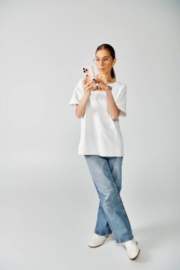 Young woman in white t-shirt and glasses taking a picture with her cell phone on a grey background. clipart