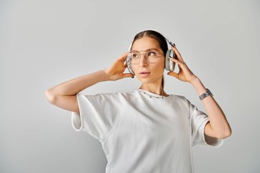 Gözlüklü bir kadın kulağına kulaklık dayamış. Gri bir arka planda beyaz tişörtle zeka ve entelektüellik yayıyor..