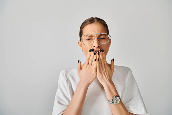 A young woman in a white t-shirt and glasses covering her mouth with her hands on a grey background.