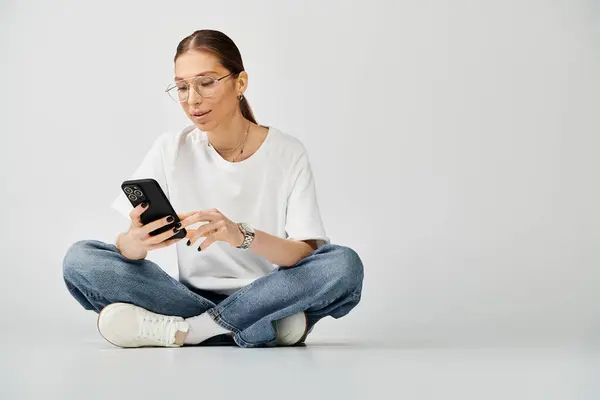 Młoda Kobieta Białej Koszulce Okularach Siedzi Podłodze Pochłonięta Telefonem Komórkowym — Zdjęcie stockowe