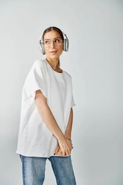 白いTシャツの若い女性は 穏やかさを誇るグレーの背景に対してヘッドフォンを通して音楽に耳を傾ける — ストック写真