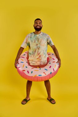 A man joyfully holds a colorful donut-shaped inflatable ring, contrasting against a sunny blue sky, embodying relaxation and fun by the water. clipart