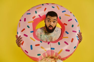 Bir adam neşe içinde kocaman pembe bir donut tutar. Renkli şekerlerle kaplıdır. Tuhaf bir zevk ve hoşgörü anı sergiler..
