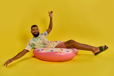 Bir adam canlı pembe bir donut şeklindeki yastığın üzerinde rahat bir şekilde oturur, kaprisli ve eğlenceli bir sahne sergiler..
