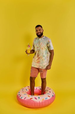 Günlük kıyafetli bir adam renkli bir donut şeklindeki bir havuzda duruyor, elinde bir içki tutuyor ve anın tadını çıkarıyor..