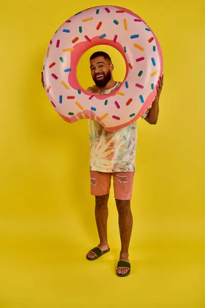 遊び心のある男が顔の前に巨大なドーナツを持ち それを完全に覆っている 彼の喜びの表現とカラフルなスパイクコントラスト — ストック写真