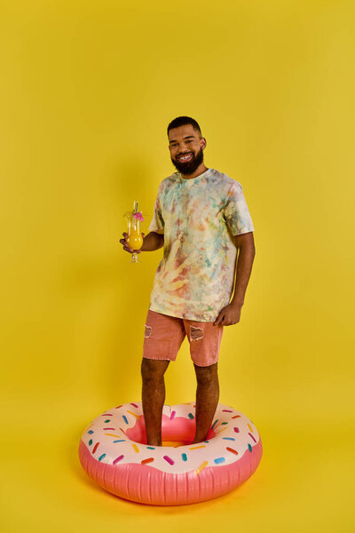 A man in casual attire stands on a colorful donut-shaped float in a pool, holding a drink in his hand and enjoying the moment.