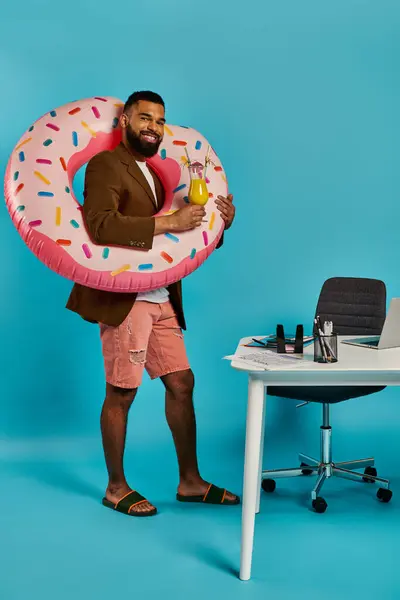 stock image A man with a playful smile holds a large inflatable donut in front of a cluttered desk, creating a whimsical and surreal scene.