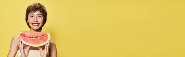 Молодая женщина игриво балансирует кусок арбуза на лице на ярком фоне.