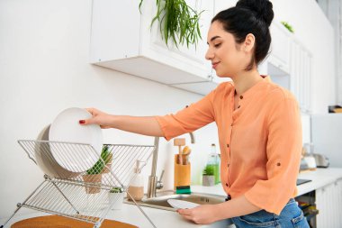 Günlük kıyafetli bir kadın mutfağında bulaşık yıkıyor, odaklanmış ve sakin..
