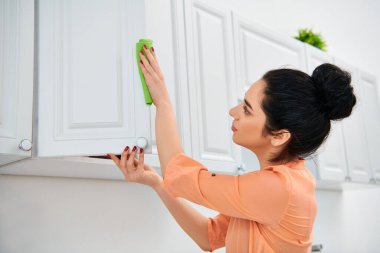 A woman in casual attire meticulously cleans a kitchen using a green rag, ensuring every surface shines with a sparkle. clipart
