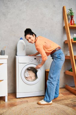 Günlük kıyafetleri içinde şık bir kadın çamaşır makinesinin yanında durmuş, evde kıyafetlerini temizlemeye odaklanmış..