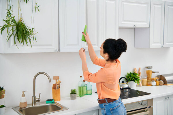 A stylish woman in casual attire methodically scrubs the kitchen sink with a vibrant green rag, bringing radiant cleanliness.