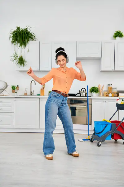 キッチンで喜んで踊るスタイリッシュな女性は 彼女の家を掃除しながらヘッドフォンを着ています — ストック写真