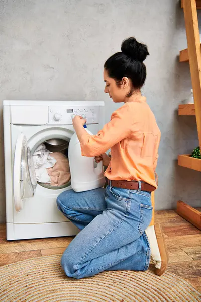 Stock image A stylish woman in casual attire sits on the floor beside a washing machine, preparing to do laundry.