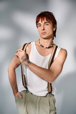 Tarz sahibi, kızıl saçlı genç bir adam beyaz bir gömlek ve pantolon askısı ile gri bir stüdyo arkaplanına poz veriyor..