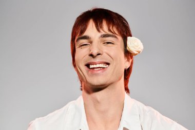 A young man striking a pose with a vibrant flower tucked behind his ear, in a studio setting against a grey background. clipart