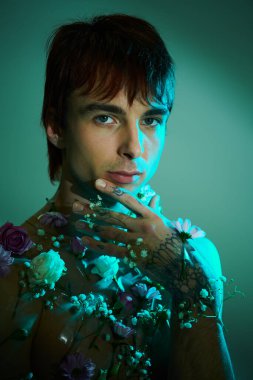 Genç bir adam mavi bir arkaplanda göğsünü renkli dövmelerle ve canlı çiçeklerle donatır..