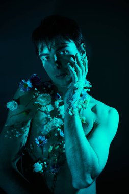 Çiçeklerle çevrili bir stüdyoda üstsüz genç bir adam erkeklik ve yumuşaklık karışımı sergiliyor..