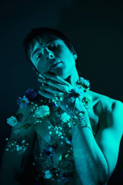 Genç bir adam bir stüdyoda boynunda çiçeklerle üstsüz duruyor.