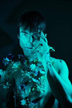 Stüdyoda mavi ışıkla poz veren çiçeklerle çevrili üstsüz genç bir adam.