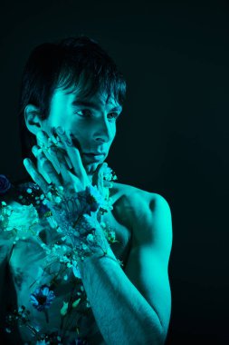 Stüdyoda mavi ışıkla poz veren çiçeklerle çevrili üstsüz bir adam.
