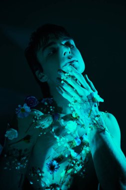 Elleri ağzında, etrafı çiçeklerle, mavi ışıkla çevrili genç bir adam.
