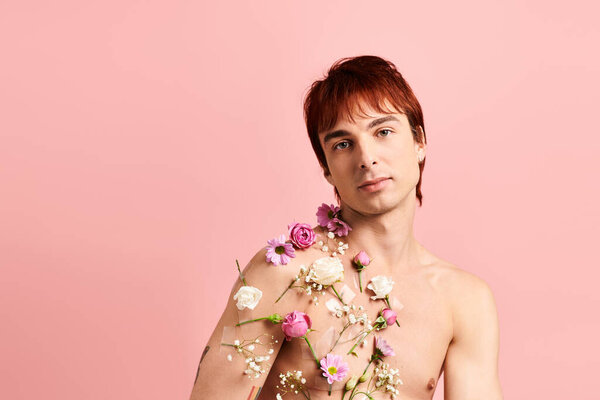 A shirtless young man poses confidently with vibrant flowers adorning his chest in a studio setting with a pink background