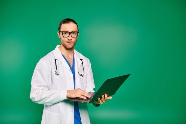 Laboratuvar önlüklü yakışıklı bir doktor yeşil bir arkaplanın önünde kendine güvenen bir laptop tutuyor..