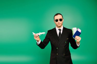 Handsome pilot in black suit poses with tickets and passport and model airplane. clipart