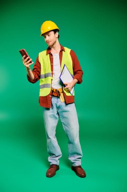 Sarı güvenlik yeleği giyen yakışıklı bir erkek işçi cep telefonunu kendine güvenerek tutuyor..