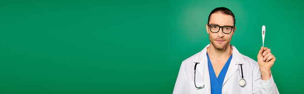 Handsome doctor in white coat holding a thermometer against a green backdrop.
