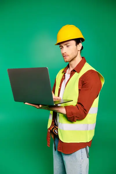 stock image Handsome worker in hard hat holding a laptop against a green backdrop.