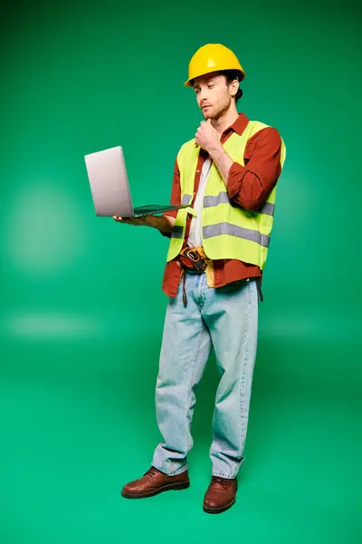 Handsome worker in hard hat holding laptop on green backdrop.