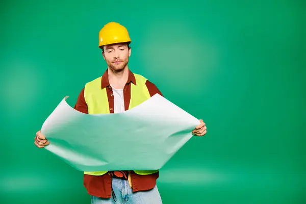 A handsome male worker in a hard hat holds a paper on a green backdrop.
