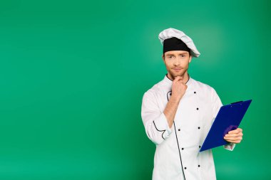 Handsome male chef in white uniform holding a clipboard on green backdrop. clipart