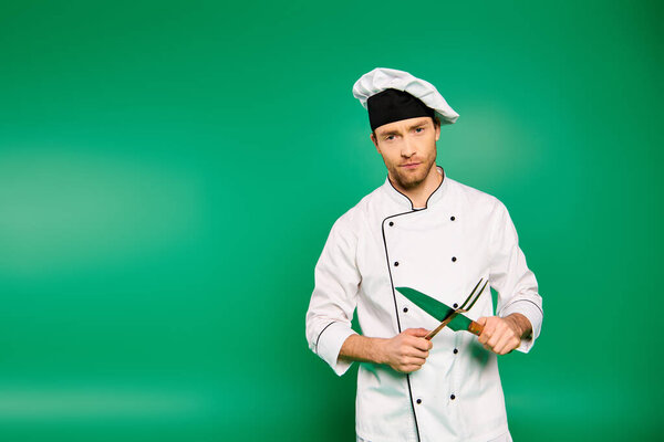 Handsome male chef in white uniform skillfully holding cutlery on green backdrop.