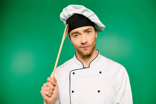 一个穿着白色制服 头戴绿色背景的英俊男性厨师 手里拿着一只木制勺子 — 图库照片